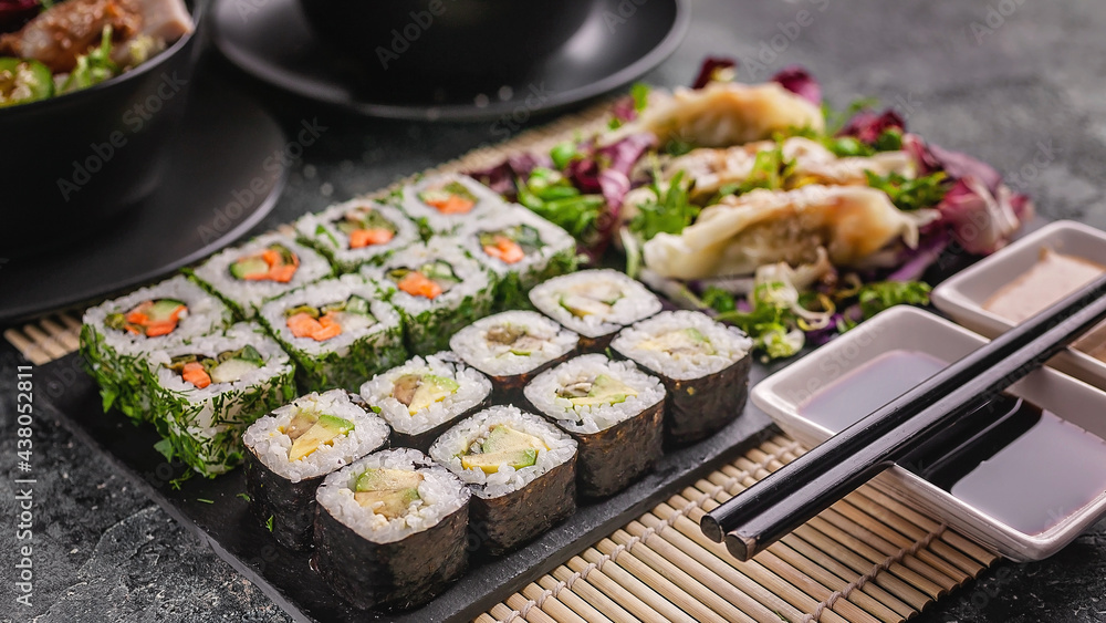 Food banner: tasty traditional Asian food. Vegetable sushi maki rolls, gyoza dumplings with salad on a black stone board. Bamboo mat, nut and soy sauces, black chopsticks. Close-up Food banner: tasty traditional Asian food. Vegetable sushi maki rolls, gyoza dumplings with salad on a black stone board. Bamboo mat, nut and soy sauces, black chopsticks. Close-up