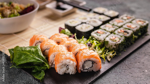 Appetizing rolls with salmon and Philadelphia cream cheese with fresh tasty salad on a black stone cutting board. Mix of traditional Asian dishes. Food banner. Copy space
