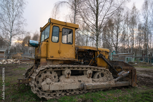 An old rusty yellow tractor among the trees in the open air on a driverless farm. Agricultural machinery