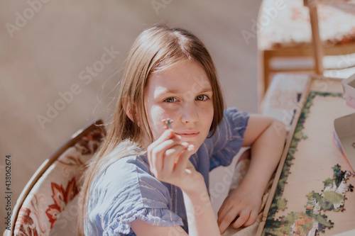 Cute little girl 8 -12 years old sitting at table holding one piece puzzle in hand and looking at camera. Brown-haired kid with long hair wearing casual blue dress.