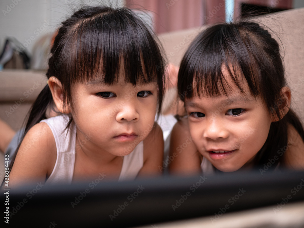 Two sibling girls lying on the sofa and watching digital tablets together in the living room. Two sibling girls lying on the sofa and watching digital tablets together in the living room.