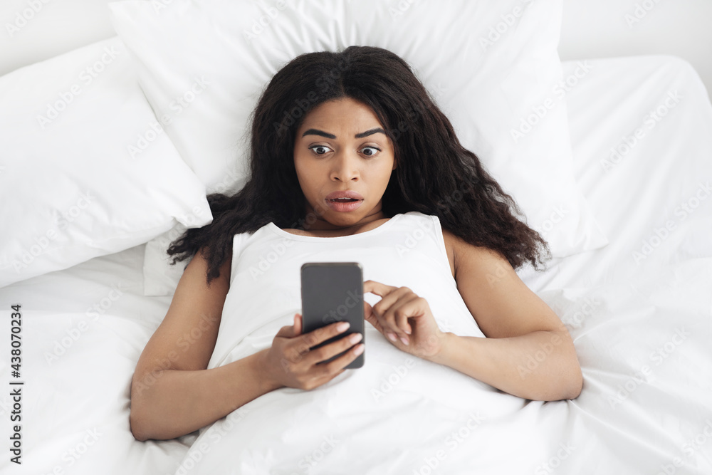 Amazing news in the morning. Shocked african american woman lying in soft bed and looking at smartphone, top view Amazing news in the morning. Shocked african american woman lying in soft bed and looking at smartphone, top view
