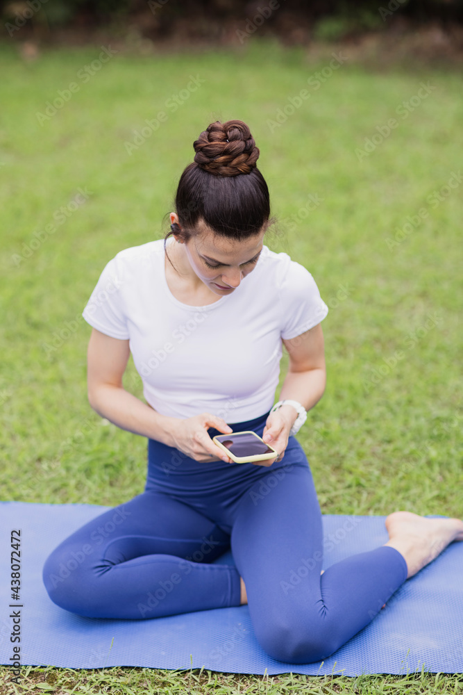Young caucasian Woman relaxing after virtual yoga class in summer park during COVID-19 coronavirus pandemic. Social distancing while fitness exercising. Girl holding mobile phone, listening music via Young caucasian Woman relaxing after virtual yoga class in summer park during COVID-19 coronavirus pandemic. Social distancing while fitness exercising. Girl holding mobile phone, listening music via