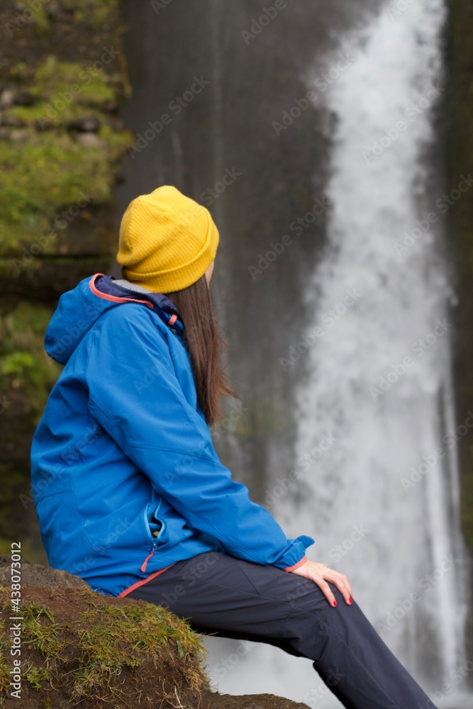 Girl with blue jacket and yellow hat from behind admiring an Icelandic waterfall Girl with blue jacket and yellow hat from behind admiring an Icelandic waterfall