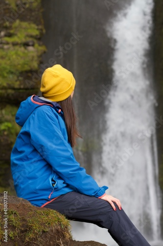 Girl with blue jacket and yellow hat from behind admiring an Icelandic waterfall