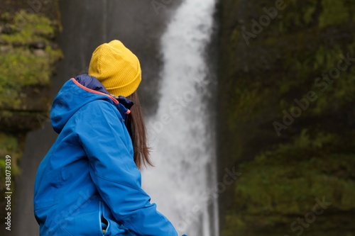 Girl with blue jacket and yellow hat from behind admiring an Icelandic waterfall © Anna