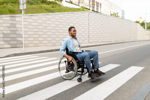 Full length portrait of impaired young black guy in wheelchair crossing city street, copy space © Prostock-studio