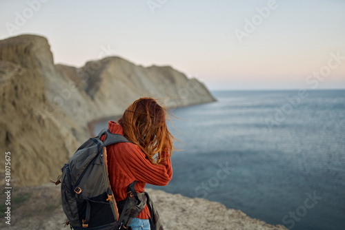 red-haired traveler with a backpack on her back looks at the sea and mountains in the distance