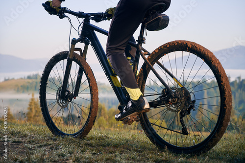 Horizontal cropped snapshot of biker's legs, sitting on bicycle. Close-up view on bike, wheels in mud, after morning ride on mountains trails. Athlete enjoying the moment of sunrise