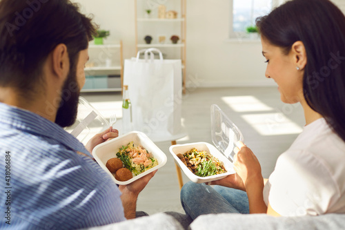Happy people enjoying takeout lunch at home. Young couple sitting on sofa and eating fresh healthy takeaway food from plastic containers. Man and woman comparing two meals ordered in delivery service photo