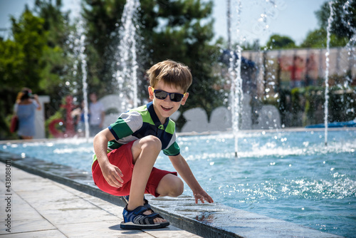 Fototapeta Naklejka Na Ścianę i Meble -  Little boy plays in the square near pool with water jets in the fountain at sunny summer day. Active summer leisure for kids in the city