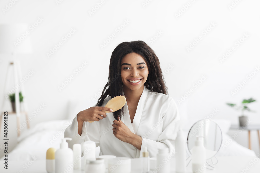 Daily hair care at home during covid-19 lockdown. Happy african american lady combing her curly hair in bedroom Daily hair care at home during covid-19 lockdown. Happy african american lady combing her curly hair in bedroom