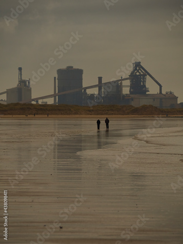 Bleached, stark landscape, a dull sky reflected on wet sand, two people silhouetted in the mid ground and a near-silhouetted steel mill on the horizon.