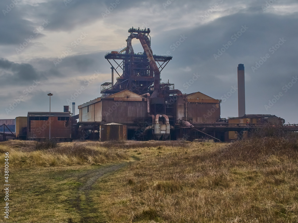 The derelict, abandoned hulk of the blast furnace at the Redcar Steelworks against a dramatic sky, seen from the grass-covered slagfields.