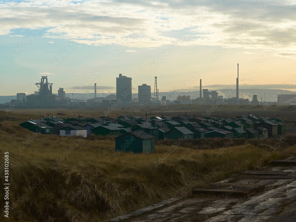 Dunes shelter a collection of small fishermans’ huts in golden grass, ahead of a silhouetted industrial horizon of the Redcar Steelworks and Teesport. Dunes shelter a collection of small fishermans’ huts in golden grass, ahead of a silhouetted industrial horizon of the Redcar Steelworks and Teesport.