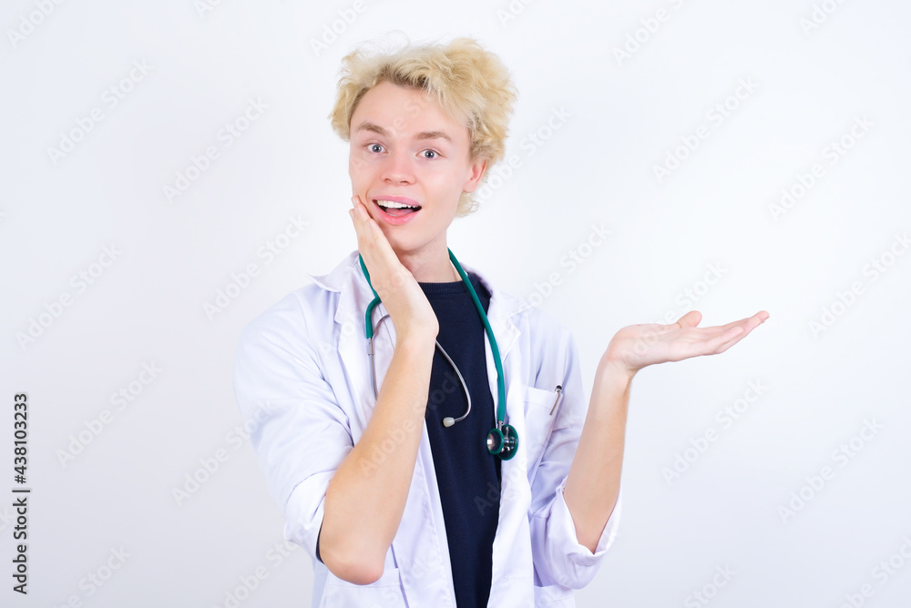 Positive glad young handsome Caucasian doctor man standing against white background says: wow how exciting it is, indicates something. One hand on his head and pointing with other hand. Positive glad young handsome Caucasian doctor man standing against white background says: wow how exciting it is, indicates something. One hand on his head and pointing with other hand.