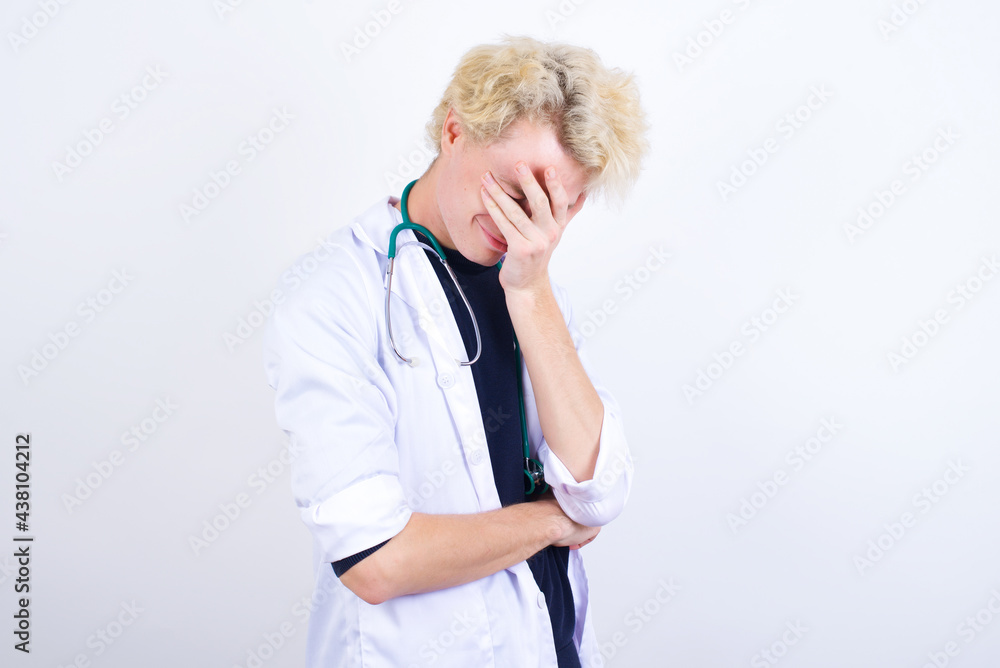 young handsome Caucasian doctor man standing against white background making facepalm gesture while smiling amazed with stupid situation. young handsome Caucasian doctor man standing against white background making facepalm gesture while smiling amazed with stupid situation.
