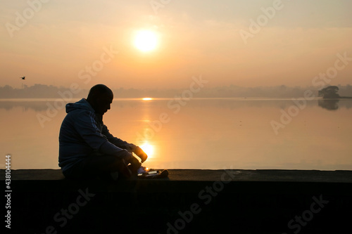 Stock photo of 50 to 60 aged Old Indian man sitting alone near lake and enjoying beautiful clam sunrise at Kolhapur Maharashtra India.