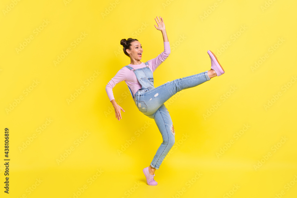 Profile photo of girl stand tiptoe dance raise hand open mouth wear denim overall sneakers isolated yellow background