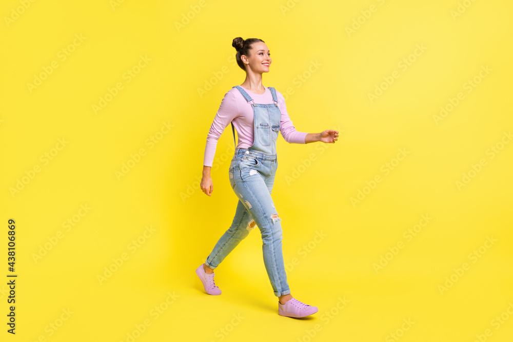 Full length photo of adorable sweet young lady dressed jeans overall walking looking empty space isolated yellow color background