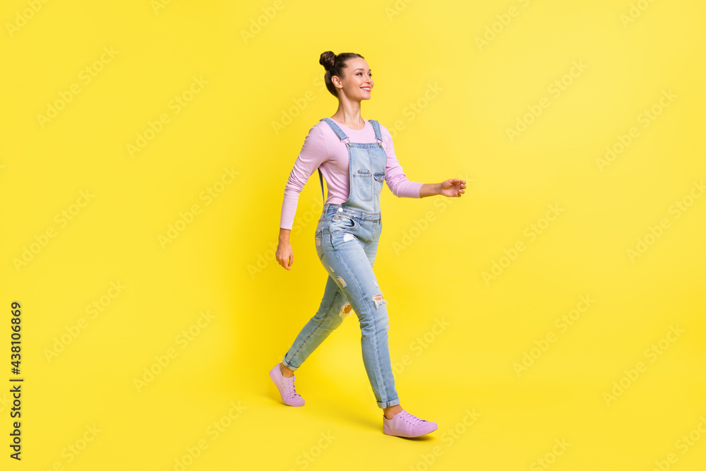 Full length photo of adorable sweet young lady dressed jeans overall walking looking empty space isolated yellow color background Full length photo of adorable sweet young lady dressed jeans overall walking looking empty space isolated yellow color background