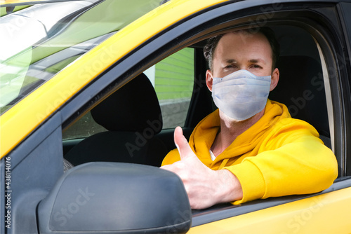 Young happy taxi driver in protective mask sits behind the wheel of a taxi