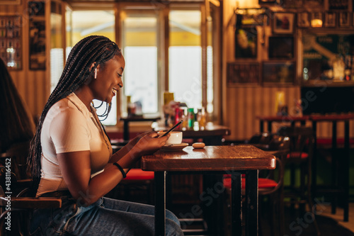 Cheerful african american woman using a smartphone and airpods while sitting in a cafe