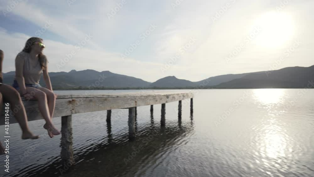 Multiracial happy gen z friends group sitting with dangling feet on a pier at sunset or dawn looking each other. Camera pan on sea ocean or flat lake water with sun reflection effect. People on jetty Multiracial happy gen z friends group sitting with dangling feet on a pier at sunset or dawn looking each other. Camera pan on sea ocean or flat lake water with sun reflection effect. People on jetty