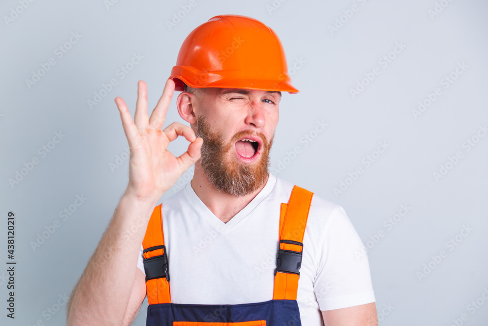 Handsome man engineer in building protective helmet on gray background shows OK gesture blinking eye Handsome man engineer in building protective helmet on gray background shows OK gesture blinking eye