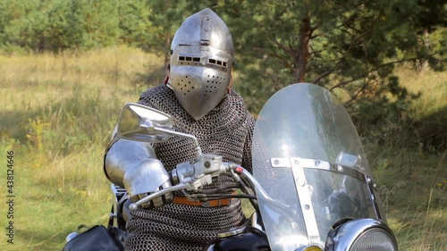 A medieval warrior in chain mail and a helmet sits at the wheel of a motorcycle against the backdrop of the forest. Biker knight concept.