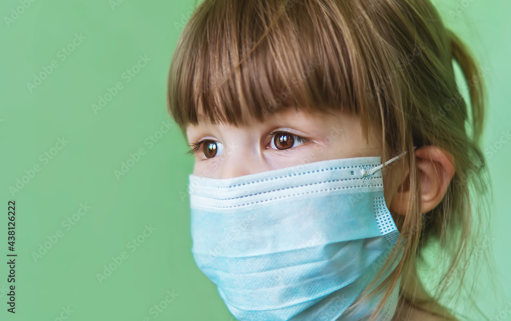 A sad little girl in a medical mask on a green background. Disposable medical mask on the child's face.A child in a protective medical mask during an epidemic.Protection against viruses and infections A sad little girl in a medical mask on a green background. Disposable medical mask on the child's face.A child in a protective medical mask during an epidemic.Protection against viruses and infections