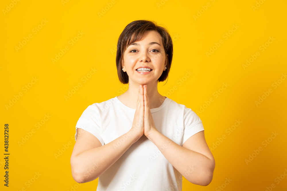 Hopeful pretty female presses palms together in pray, believes in wellness, has big faith, wears casual t-shirt, stays calm, isolated over yellow background. May my dreams come true. Hopeful pretty female presses palms together in pray, believes in wellness, has big faith, wears casual t-shirt, stays calm, isolated over yellow background. May my dreams come true.