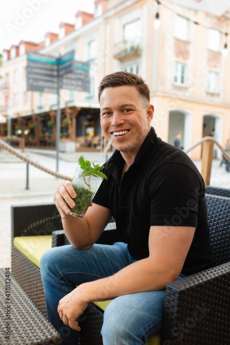 Young man sitting at table of street cafe with cooling mojito cocktail, smiling and looking at camera