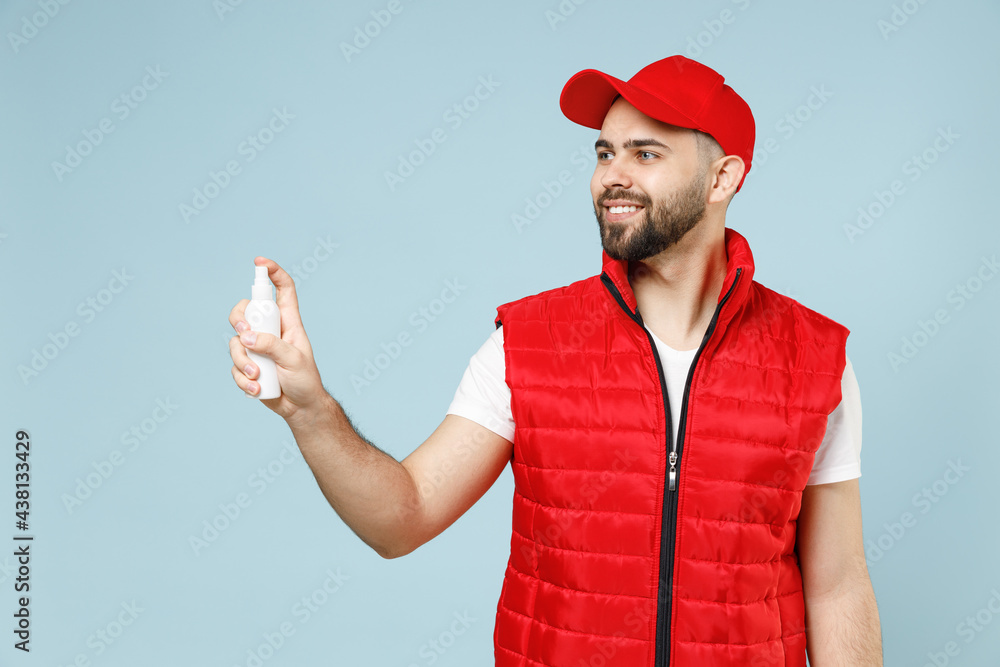 Delivery guy employee man in red cap white T-shirt vest uniform workwear work dealer courier hold bottle spray sanitizer soap isolated on pastel blue color background studio portrait. Service concept. Delivery guy employee man in red cap white T-shirt vest uniform workwear work dealer courier hold bottle spray sanitizer soap isolated on pastel blue color background studio portrait. Service concept.