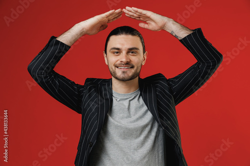 Young fun businessman latin man long hair ponytail wear black striped jacket grey shirt hold folded hands above head like house roof, stay home isolated on red background. Concept of people lifestyle.