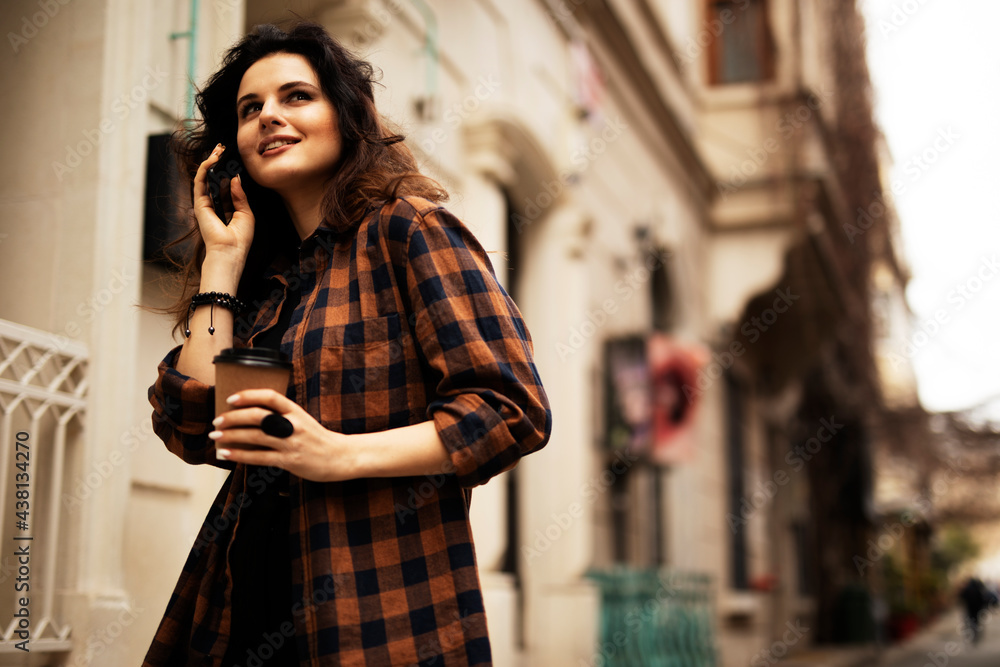 Young smiling woman drinking coffee outdoors. Beautiful woman using the phone while walking in the morning