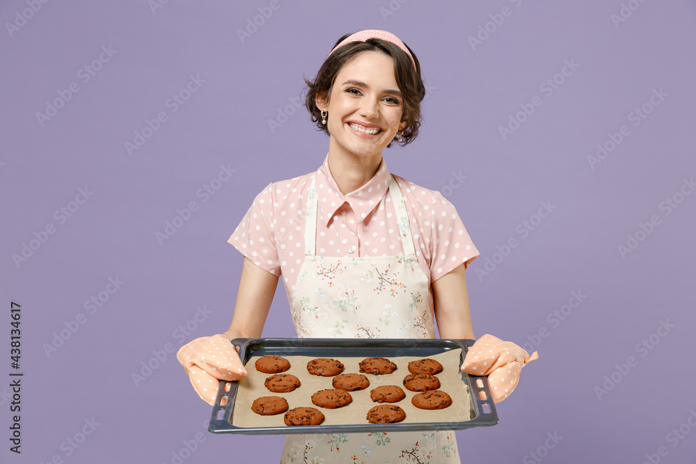 Young smiling happy housewife housekeeper chef cook baker woman wearing pink apron show chocolate cookies biscuits on baking sheet isolated on pastel violet background studio. Cooking food concept Young smiling happy housewife housekeeper chef cook baker woman wearing pink apron show chocolate cookies biscuits on baking sheet isolated on pastel violet background studio. Cooking food concept
