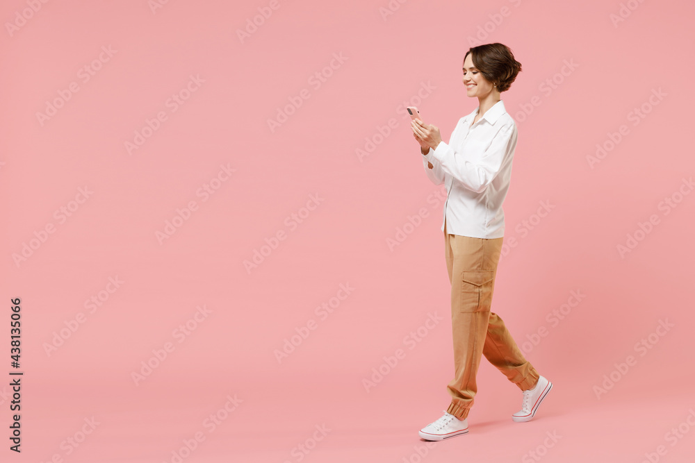 Full length side view young employee business woman corporate lawyer in classic formal white shirt work in office hold using mobile cell phone chat online isolated on pastel pink background studio Full length side view young employee business woman corporate lawyer in classic formal white shirt work in office hold using mobile cell phone chat online isolated on pastel pink background studio