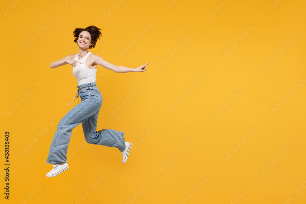 Full length young fun happy excited woman 20s with bob haircut wearing white tank top shirt jumping high point index finger aside on copy space area mock up workspace isolated on yellow background