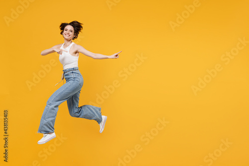 Full length young fun happy excited woman 20s with bob haircut wearing white tank top shirt jumping high point index finger aside on copy space area mock up workspace isolated on yellow background