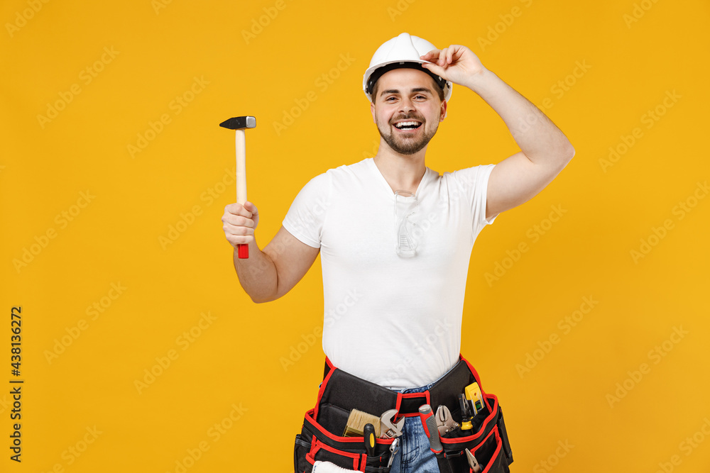 Young smiling employee handyman man in protective helmet glasses hold hammer tipping hardhat isolated on yellow background studio Instruments accessories renovation apartment room Repair home concept. Young smiling employee handyman man in protective helmet glasses hold hammer tipping hardhat isolated on yellow background studio Instruments accessories renovation apartment room Repair home concept.