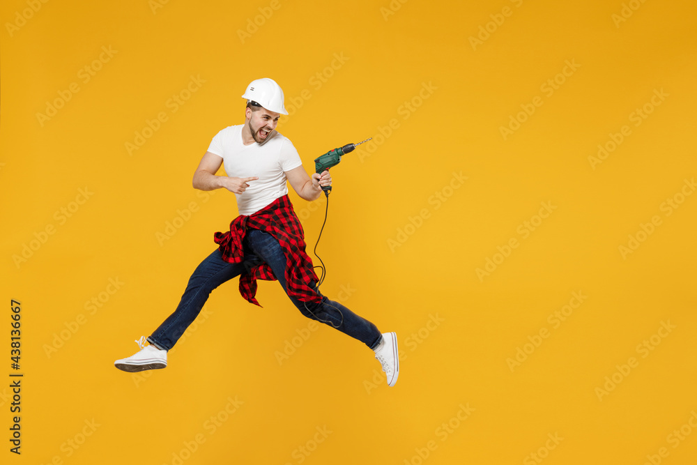 Full length young employee handyman man in t-shirt use electric drill jump point finger aside on area mock up isolated on yellow background Instruments accessories apartment room. Repair home concept.