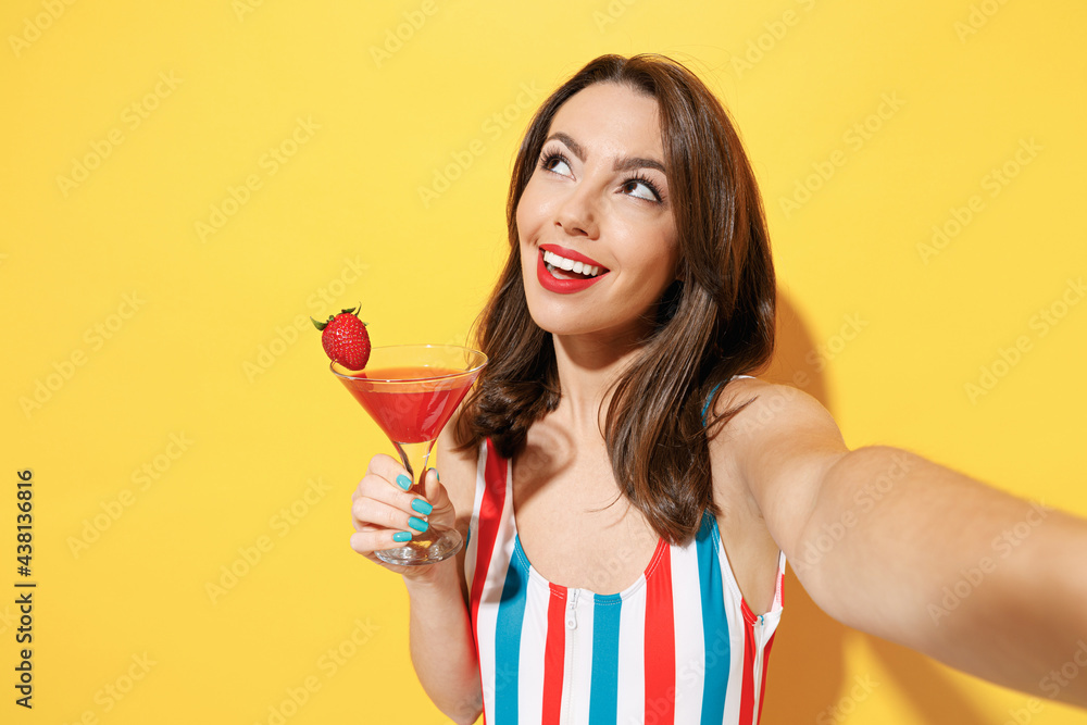 Happy young sexy woman slim body wear striped red blue one-piece swimsuit hold glass of strawberry cocktail isolated on vivid yellow color background studio Summer hotel pool sea rest sun tan concept Happy young sexy woman slim body wear striped red blue one-piece swimsuit hold glass of strawberry cocktail isolated on vivid yellow color background studio Summer hotel pool sea rest sun tan concept