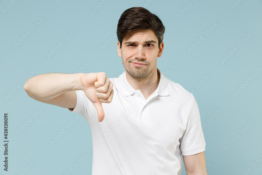 Young sad displeased disappointed unshaven caucasian man in white casual basic t-shirt showing thumb down dislike gesture isolated on pastel blue background studio portrait. People lifestyle concept Young sad displeased disappointed unshaven caucasian man in white casual basic t-shirt showing thumb down dislike gesture isolated on pastel blue background studio portrait. People lifestyle concept