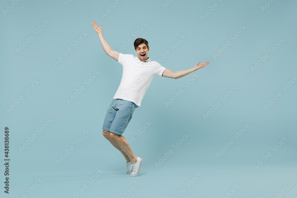 Full length young fun man 20s wearing white basic t-shirt stand with outstretched hands leaning back stand on toes isolated on pastel blue color background studio portrait. People lifestyle concept.