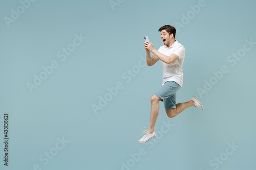 Full length side view smiling young man 20s wearing white casual basic t-shirt using mobile cell phone gesture jump high isolated on pastel blue background studio portrait. People lifestyle concept.