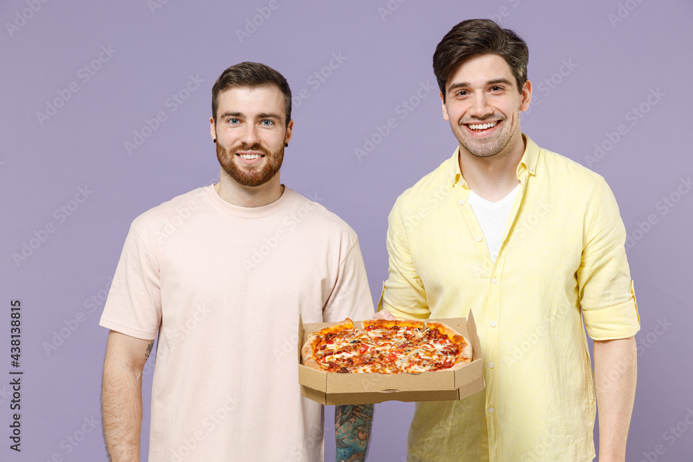 Two smiling happy cool satisifed young men friends together in casual yellow t-shirt hold italian pizza in cardboard flatbox look camera isolated on purple background studio People lifestyle concept. Two smiling happy cool satisifed young men friends together in casual yellow t-shirt hold italian pizza in cardboard flatbox look camera isolated on purple background studio People lifestyle concept.
