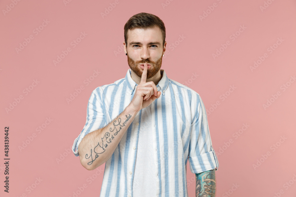 Young secret caucasian unshaven man 20s in blue striped shirt white t-shirt say hush be quiet with finger on lips shhh gesture isolated on pastel pink background studio portrait. Tattoo translate fun Young secret caucasian unshaven man 20s in blue striped shirt white t-shirt say hush be quiet with finger on lips shhh gesture isolated on pastel pink background studio portrait. Tattoo translate fun