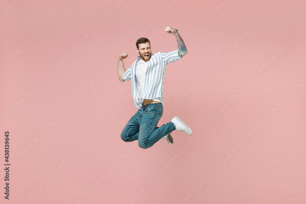 Full length young overjoyed unshaven man 20s in blue striped shirt white t-shirt jump high do winner gesture clench fist isolated on pastel pink background studio portrait. People lifestyle concept. Full length young overjoyed unshaven man 20s in blue striped shirt white t-shirt jump high do winner gesture clench fist isolated on pastel pink background studio portrait. People lifestyle concept.