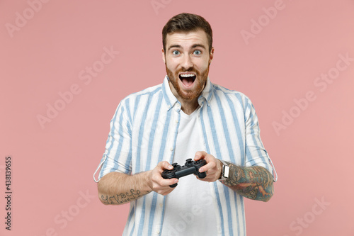 Young smiling excited happy caucasian unshaven man 20s wearing blue striped shirt playing pc game with joystick console isolated on pastel pink color background studio portrait. Tattoo translate fun